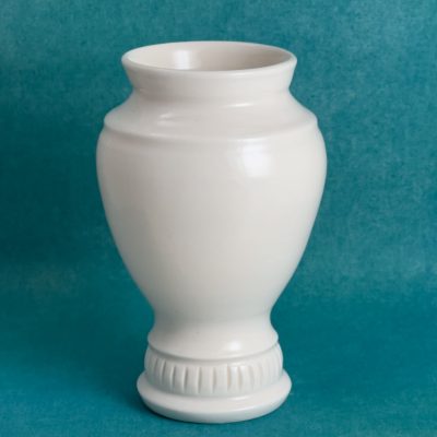 Vase en porcelaine blanche en forme d'urne, corps ovale posé sur un pied étroit orné de petites lignes verticales, sur fond turquoise.