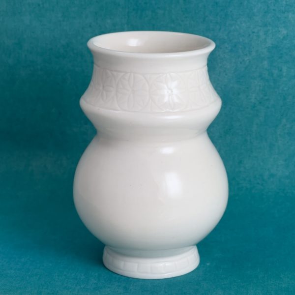 Vase à bande fleurie Vase en porcelaine blanche avec une bande à motif de fleurs autour du col, un corps rond et un petit pied lui aussi décoré, sur fond turquoise.