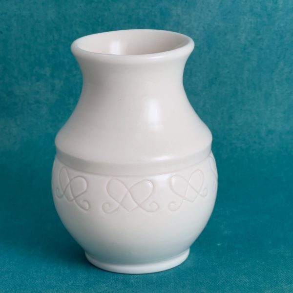 Vase en porcelaine blanche avec un col évasé et une bande de petits motifs en forme de cœurs reliés autour du bas du corps, sur fond turquoise.
