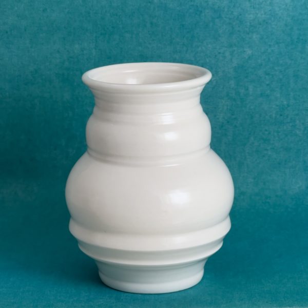 Vase à large ouverture Vase en porcelaine blanche à large ouverture évasée, col court et panse ronde marquée par plusieurs lignes horizontales, sur fond turquoise.