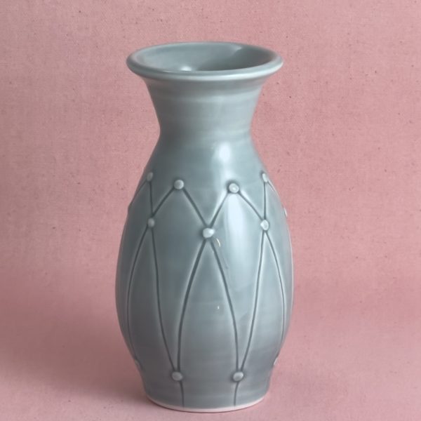 Vase en porcelaine gris-bleu au col évasé, dont la panse est ornée d'un treillis en losanges gravé, ponctué de petites perles appliquées à chaque intersection, photographié sur fond de lin rose.