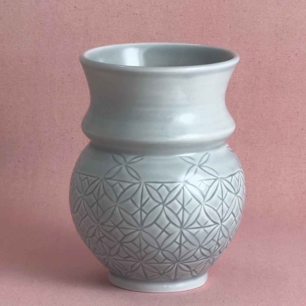 Vase en porcelaine gris-bleu au col cintré et évasé, dont la panse ronde est entièrement gravée d'un motif géométrique serré en étoiles et pétales, photographié sur fond de lin rose.
