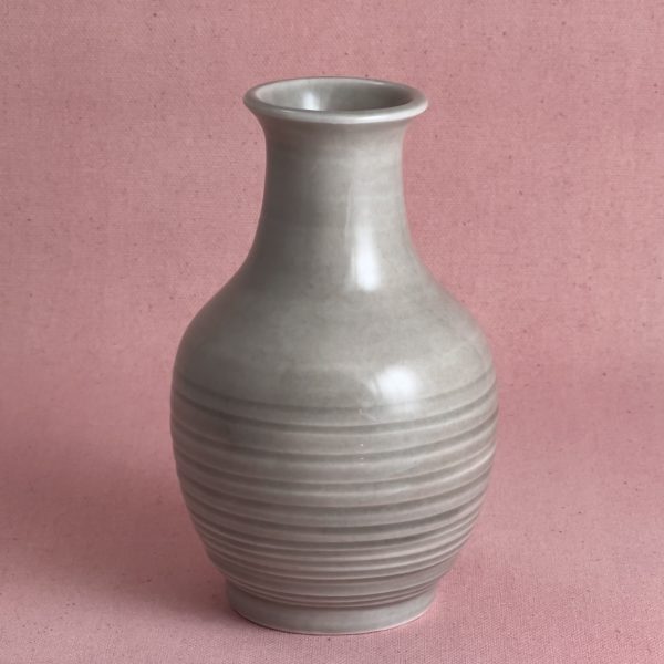 Vase en porcelaine grise en forme de bouteille, au col étroit et à l'épaule arrondie, dont la moitié inférieure est striée de fines cannelures horizontales, photographié sur fond de lin rose.