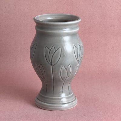 Vase gris aux tulipes Vase en porcelaine grise sur pied évasé, au col cerclé et à la panse ornée de tulipes gravées aux tiges filantes, photographié sur fond de lin rose.
