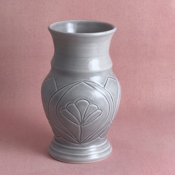 Vase gris à la fleur gravée Vase en porcelaine grise au col évasé et au pied cerclé, dont la panse porte une large fleur à pétales gravée, encadrée de deux pans pointus, photographié sur fond de lin rose.