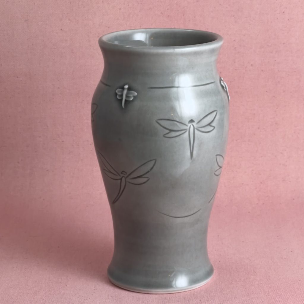 Vase en porcelaine grise au col évasé et à la panse légèrement cintrée, décoré de libellules en vol gravées et d'une libellule en relief appliquée près du col, photographié sur fond de lin rose.