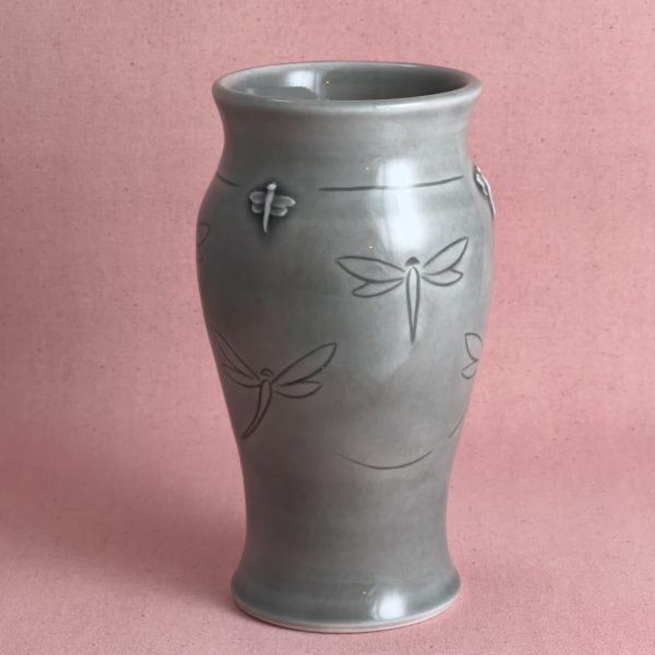 Vase gris aux libellules Vase en porcelaine grise au col évasé et à la panse légèrement cintrée, décoré de libellules en vol gravées et d'une libellule en relief appliquée près du col, photographié sur fond de lin rose.