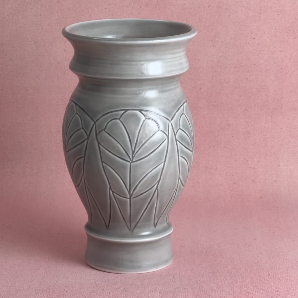Vase en porcelaine grise au col évasé et au pied cerclé, dont la panse porte de longues feuilles pointues gravées, disposées en éventail sur tout le pourtour, photographié sur fond de lin rose.