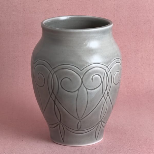 Vase gris à arabesques Vase en porcelaine grise à col évasé, orné d'un motif de volutes et d'arabesques gravées sur la panse, photographié sur fond de lin rose.