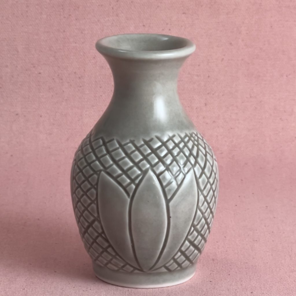 Petit vase en porcelaine grise au col évasé, dont la panse est couverte d'un quadrillage gravé en diagonale, avec un motif de pétales au centre, photographié sur fond de lin rose.