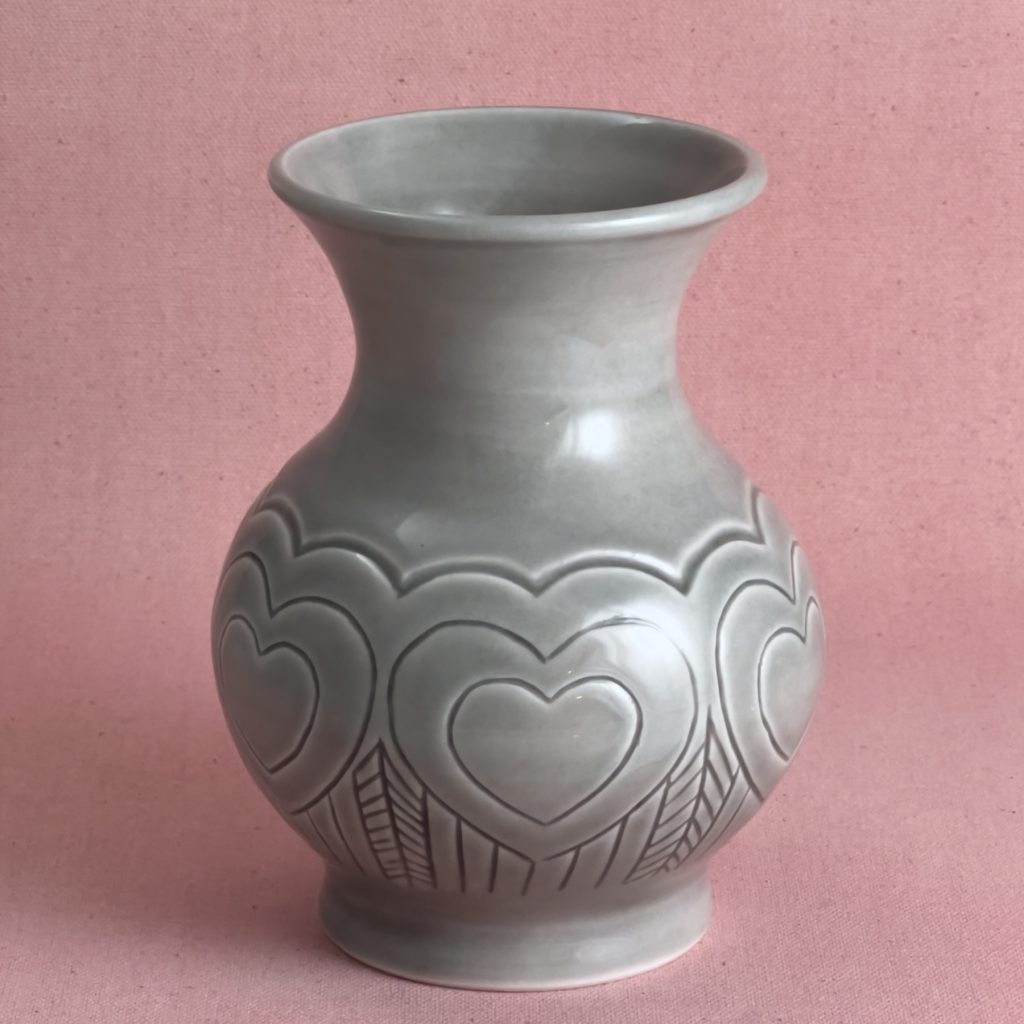 Vase en porcelaine grise au col évasé en trompette, orné d'une guirlande de cœurs gravés autour de la panse arrondie, avec des franges verticales sous chaque cœur, photographié sur fond de lin rose.